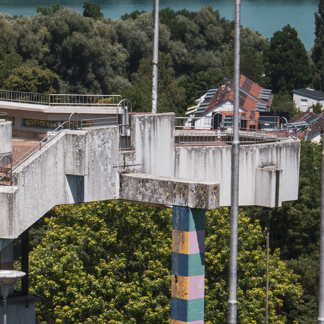 Terrasse K7 an der Universität Konstanz. Im Vordergrund eine begrünte Betonbrüstung, dahinter Bäume und Sträucher; im Hintergrund sind unscharf in der Ferne Gebäude zu sehen.