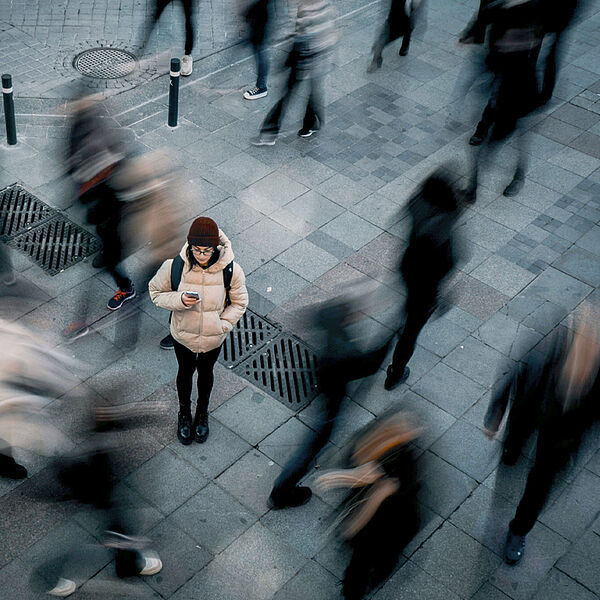 Eine Person steht auf einem gepflasterten Platz und schaut auf ihr Smartphone, während viele andere Menschen unscharf und in Bewegung um sie herum vorbeigehen.