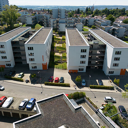 Luftaufnahme der Wohnanlage West I mit zwei parallelen vierstöckigen Gebäuden, zwischen denen sich ein begrünter Innenhof befindet. Im Vordergrund sind Stellplätze und die Jacob-Burckhardt-Straße zu sehen, im Hintergrund die Wohnanlage West II mit weiteren Grünflächen und Bäumen.