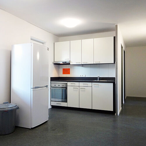 Helle Gemeinschaftsküche mit Kühlschrank, Herd, Spüle und weißen Küchenschränken und einem großen schwarzen Regal in einem Seezeit-Wohnheim