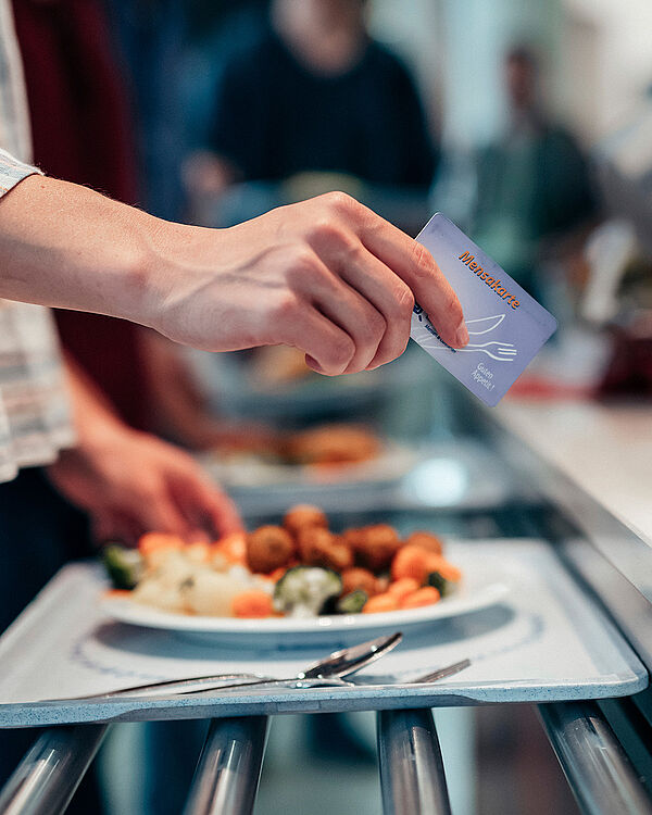 Hand hält eine Mensakarte über einem Tablett mit Essen und Besteck an einer Mensa-Ausgabe.