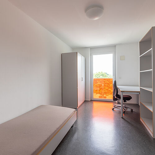 Helles Zimmer mit einem Bett links, einem Schreibtisch mit Drehstuhl rechts, einem Regal, einem Kleiderschrank und einem Fenster in Raumhöhe