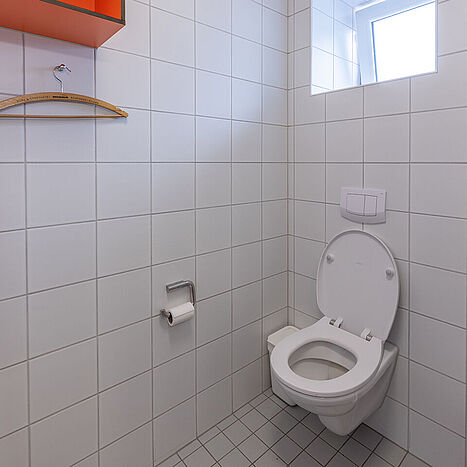 Helles Badezimmer mit Toilette, Fenster, Fliesenwänden und offenem Regal mit orangefarbenem Fach in einem Seezeit-Wohnheim