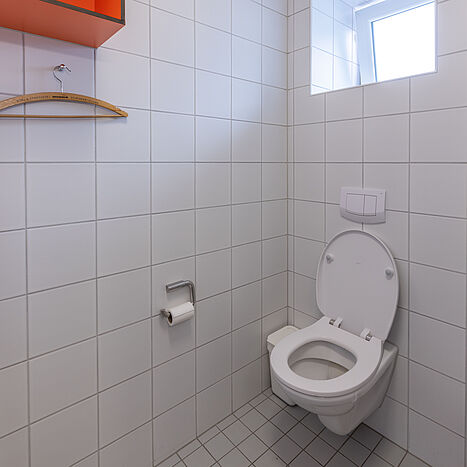 Helles Badezimmer mit Toilette, Fenster, Fliesenwänden und offenem Regal mit orangefarbenem Fach in einem Seezeit-Wohnheim