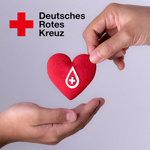 Zwei Hände, eine Hand hält ein rotes Stoffherz mit weißen Blutstropfen und einem weißen Kreuz darauf, die andere Hand formt eine Mulde zum Auffangen des Stoffherzens.