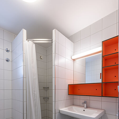 Bad in einer WG der Wohnanlage Sonnenbühl West I mit Dusche, großem Waschbecken und Modernes, orangefarbenes Badezimmerregal mit integriertem Spiegel und Lichtleiste vor weiß gefliester Wand.