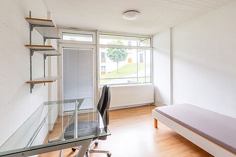 Blick vom Eingangsbereich durch das längliche Einzelzimmer zur großen Fensterfront. Vom Eingang aus steht das Bett längs an der rechten Wand; links befinden sich ein gläserner Schreibtisch mit Bürostuhl und mehrere Wandregale. Unter der Fensterfront am gegenüberliegenden Ende des Zimmers befindet sich ein Heizkörper